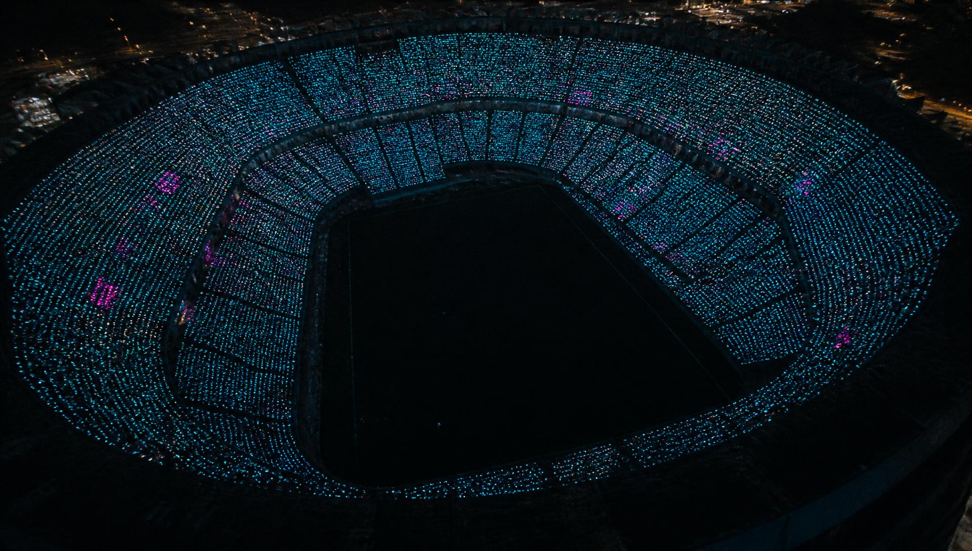 Wide aerial view of a full arena at night where thousands of illuminated jerseys form one coordinated display